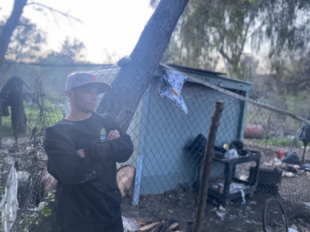 A man in a sweater and baseball cap stands at a homeless encampment in San Jose, California