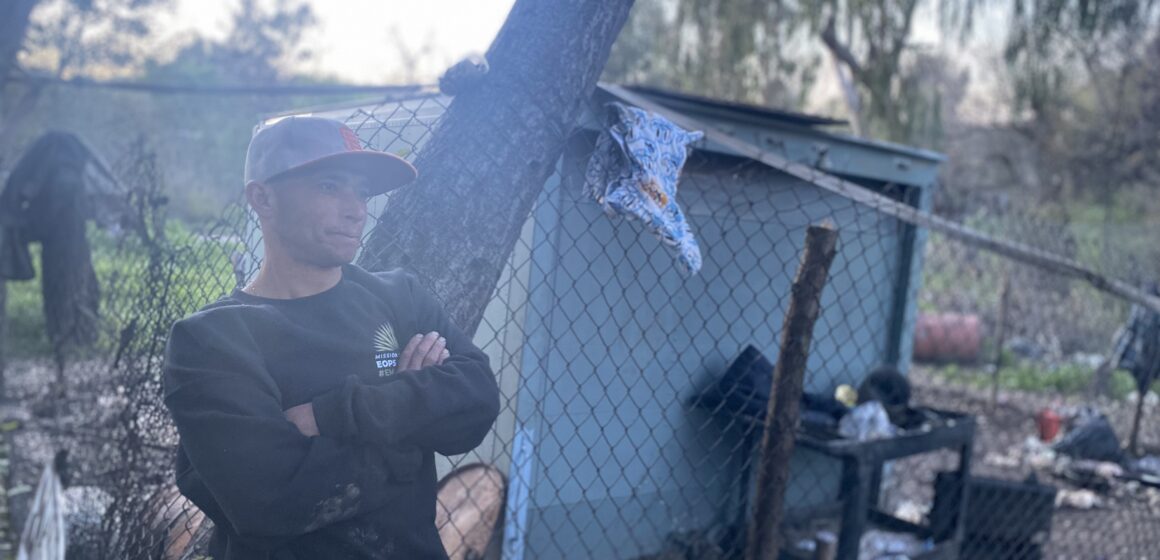 A man in a sweater and baseball cap stands at a homeless encampment in San Jose, California
