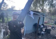 A man in a sweater and baseball cap stands at a homeless encampment in San Jose, California