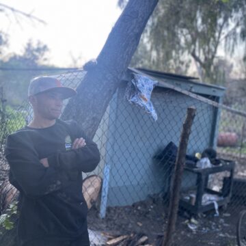 A man in a sweater and baseball cap stands at a homeless encampment in San Jose, California