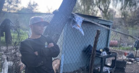 A man in a sweater and baseball cap stands at a homeless encampment in San Jose, California