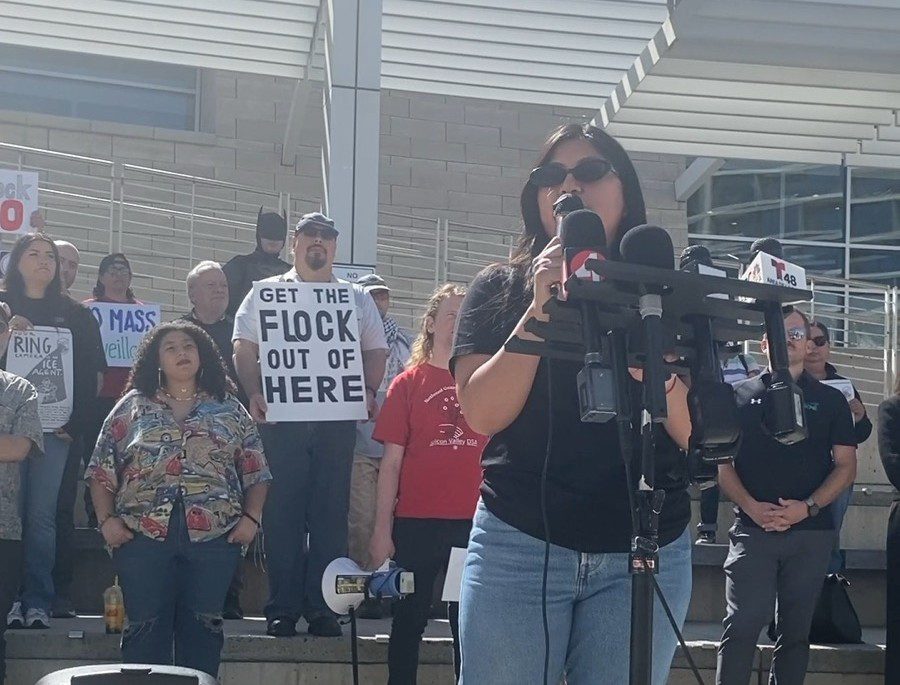 A woman speaks at a podium as demonstrators rally behind her.