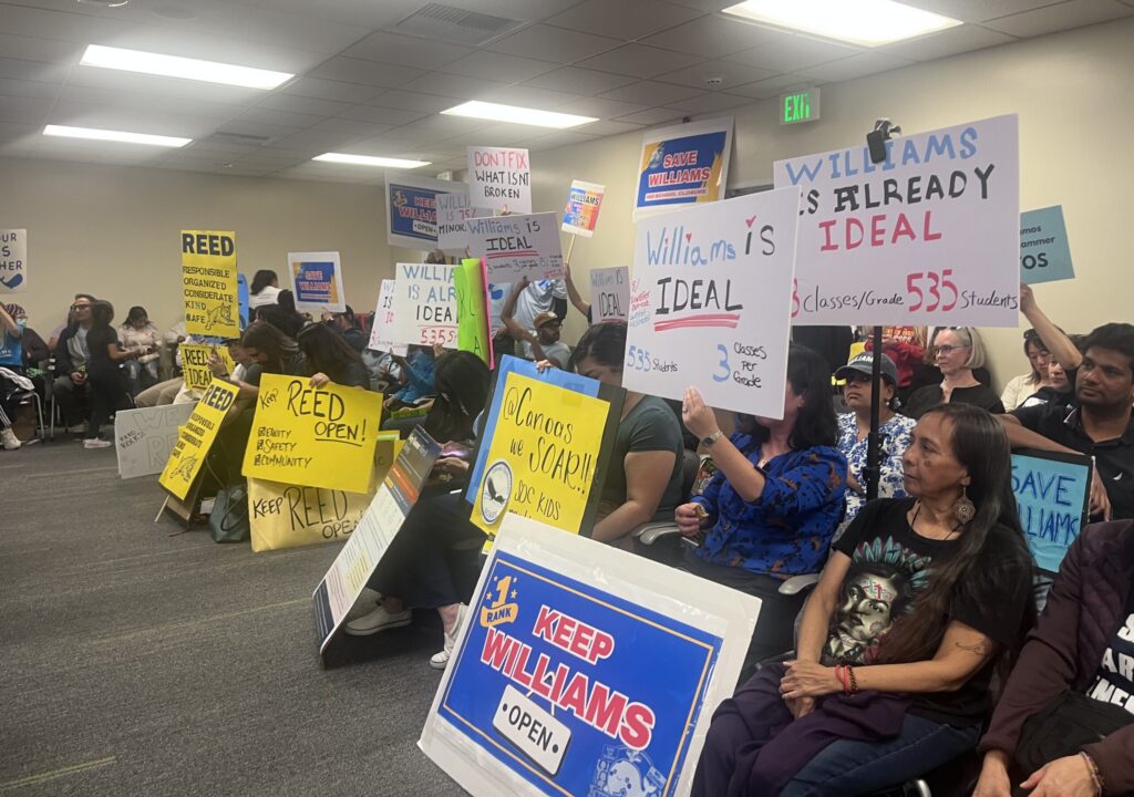 People sitting in chairs holding protest signs at a school board meeting in San Jose, California