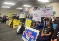 People sitting in chairs holding protest signs at a school board meeting in San Jose, California