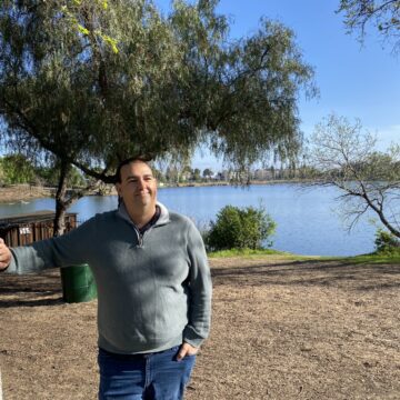 A man leans on a tree in front of a lake.