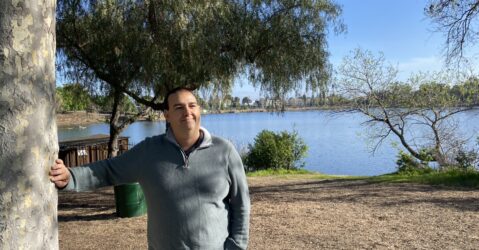 A man leans on a tree in front of a lake.