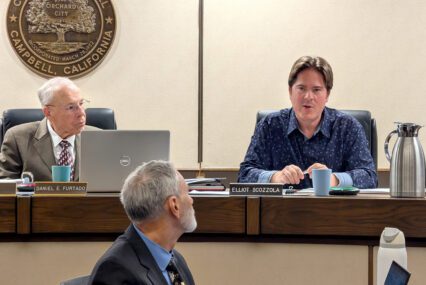 Two men up on a dais at a government meeting, with another man in front of them in Campbell, California