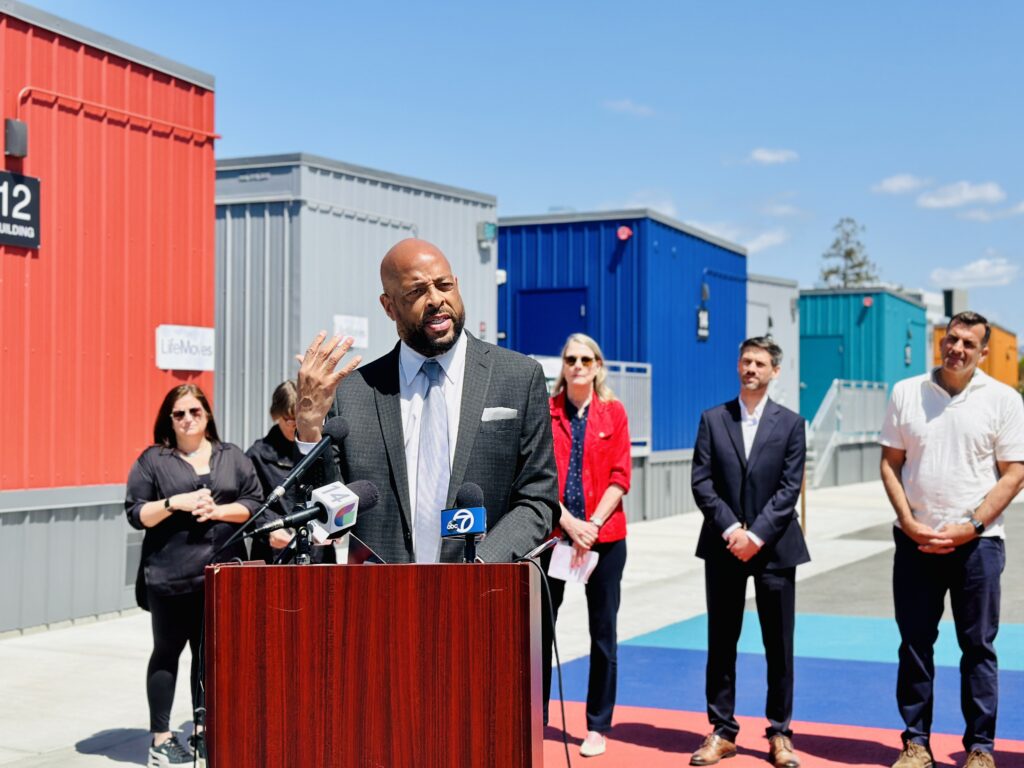 A man speaking at a podium outside, with other people standing behind him