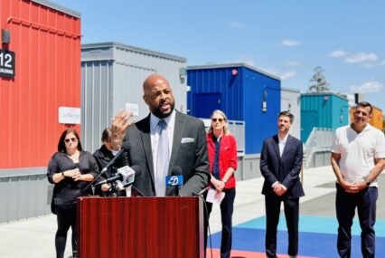A man speaking at a podium outside, with other people standing behind him