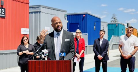 A man speaking at a podium outside, with other people standing behind him