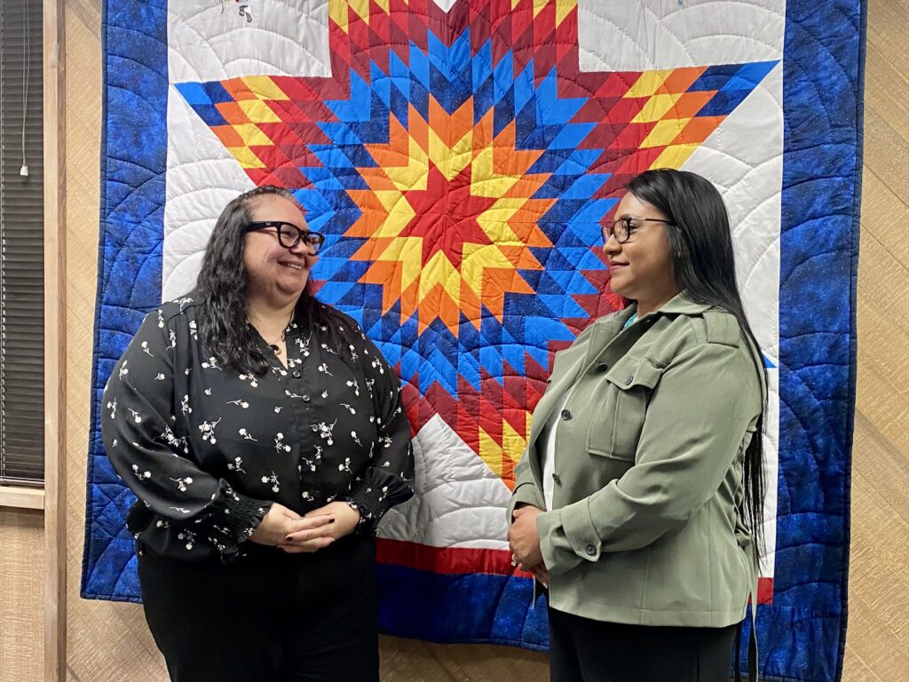 Two women stand in front of a tribal design