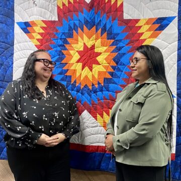 Two women stand in front of a tribal design