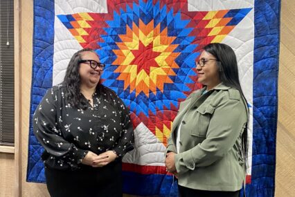 Two women stand in front of a tribal design