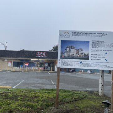 A sign on a grass field in front of a brown building next to a paved road.
