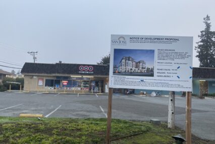 A sign on a grass field in front of a brown building next to a paved road.