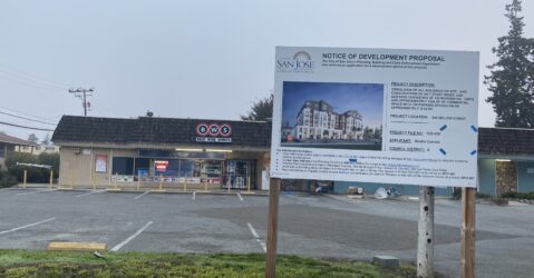 A sign on a grass field in front of a brown building next to a paved road.