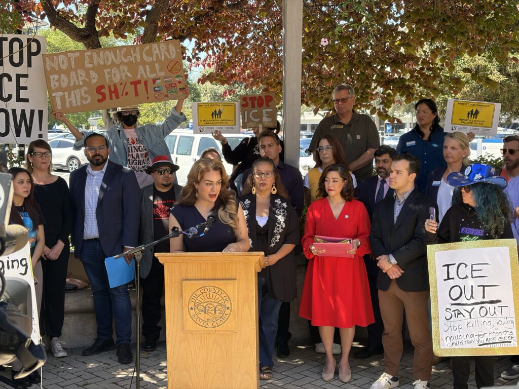 A woman speaks at a podium outside, with a crowd of people holding signs standing behind her