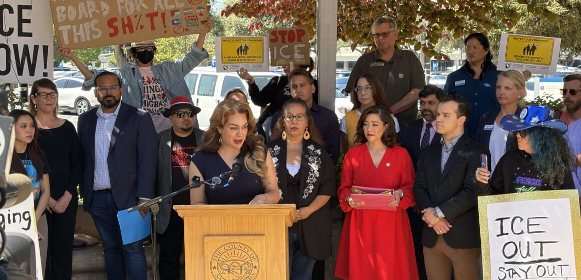 A woman speaks at a podium outside, with a crowd of people holding signs standing behind her