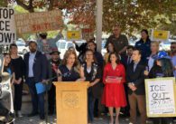 A woman speaks at a podium outside, with a crowd of people holding signs standing behind her