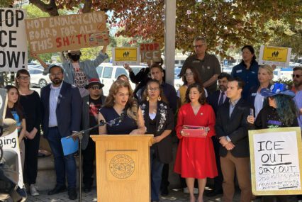 A woman speaks at a podium outside, with a crowd of people holding signs standing behind her