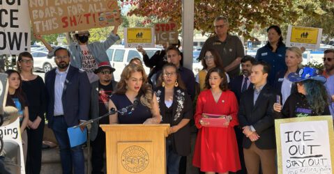 A woman speaks at a podium outside, with a crowd of people holding signs standing behind her
