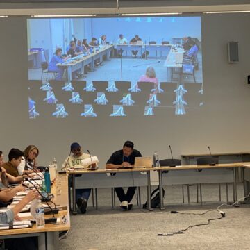 Varias personas se sientan en mesas durante una reunión gubernamental en San José, California.