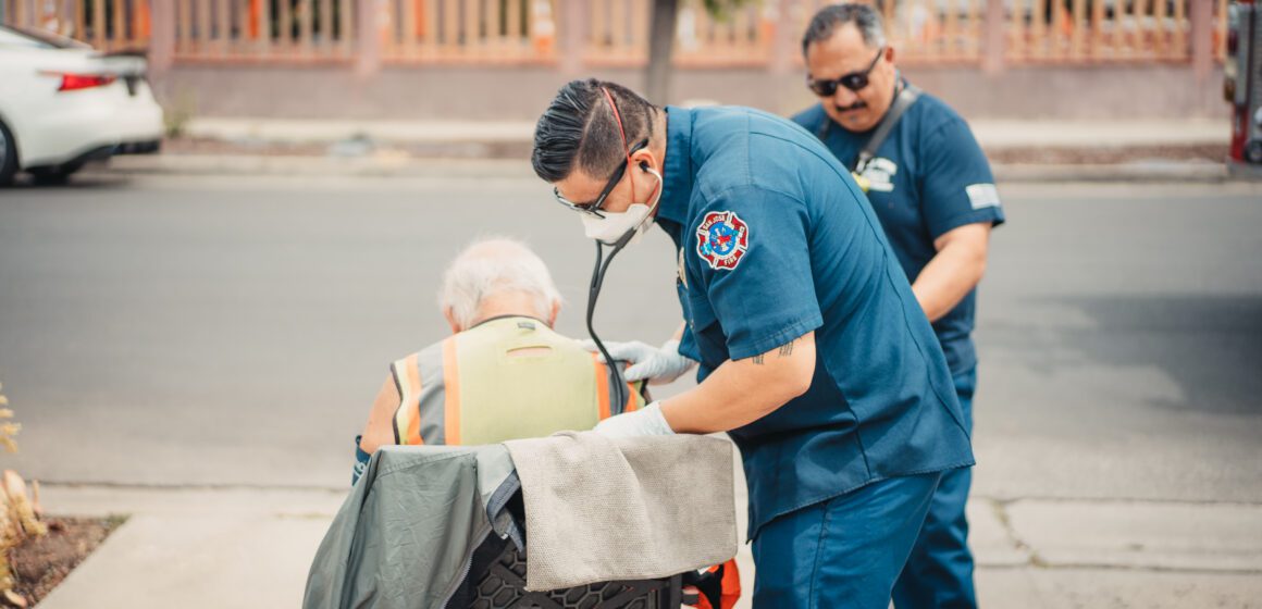 Two paramedics in blue stand next to a man in a chair.