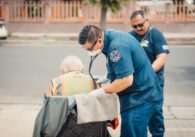 Two paramedics in blue stand next to a man in a chair.