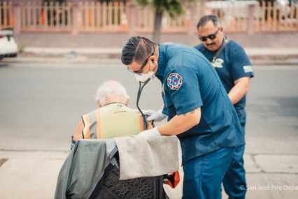 Dos paramédicos vestidos de azul están de pie junto a un hombre sentado en una silla.