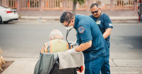 Two paramedics in blue stand next to a man in a chair.