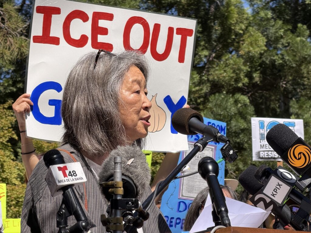 A woman stands at a podium in front of a protest sign.