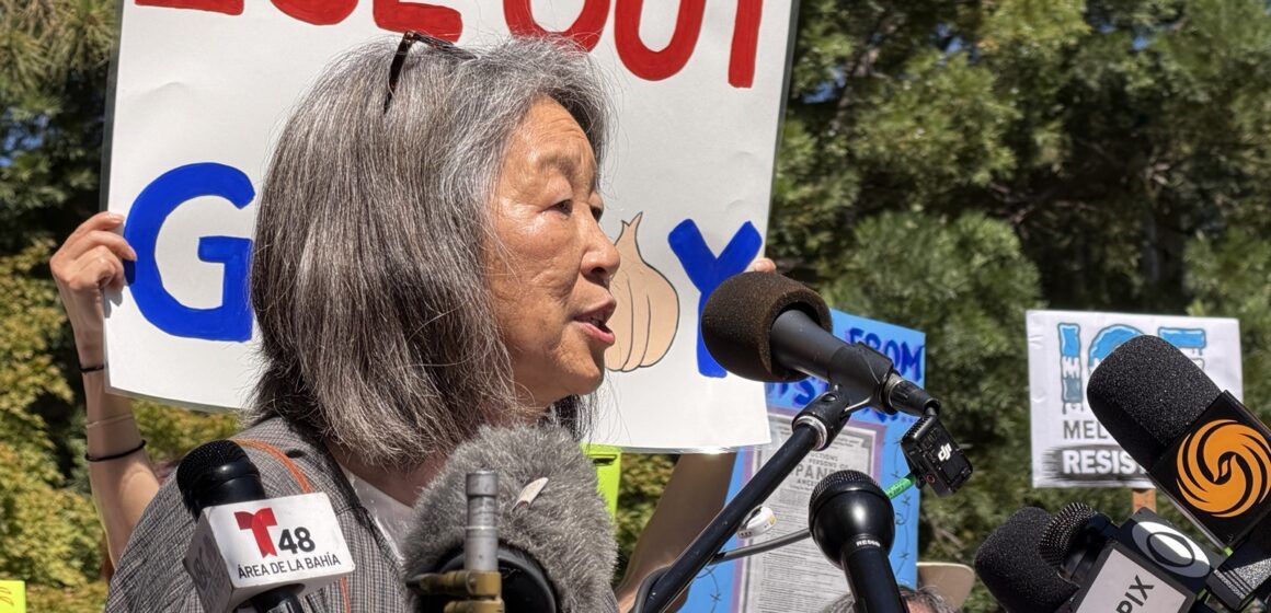 A woman stands at a podium in front of a protest sign.