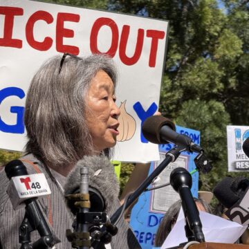 A woman stands at a podium in front of a protest sign.