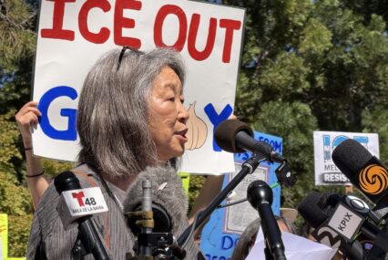 A woman stands at a podium in front of a protest sign.