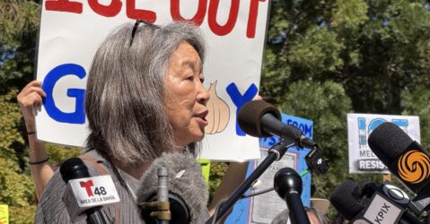 A woman stands at a podium in front of a protest sign.