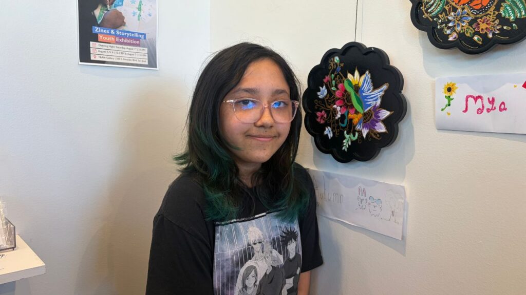 A teenager stands with artwork she painted that i hanging on a wall of an art gallery in San Jose, California