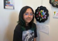 A teenager stands with artwork she painted that i hanging on a wall of an art gallery in San Jose, California