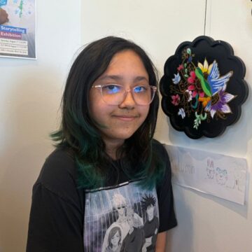 A teenager stands with artwork she painted that i hanging on a wall of an art gallery in San Jose, California