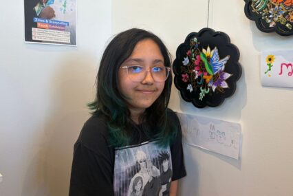 A teenager stands with artwork she painted that i hanging on a wall of an art gallery in San Jose, California