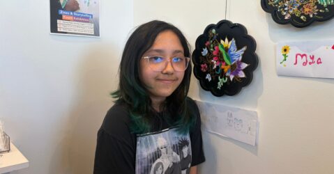A teenager stands with artwork she painted that i hanging on a wall of an art gallery in San Jose, California