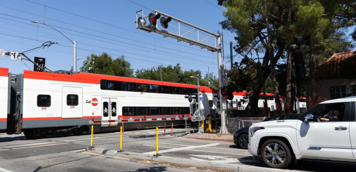 A train passes through a crossing while cars wait behind a striped barrier in Palo Alto, California