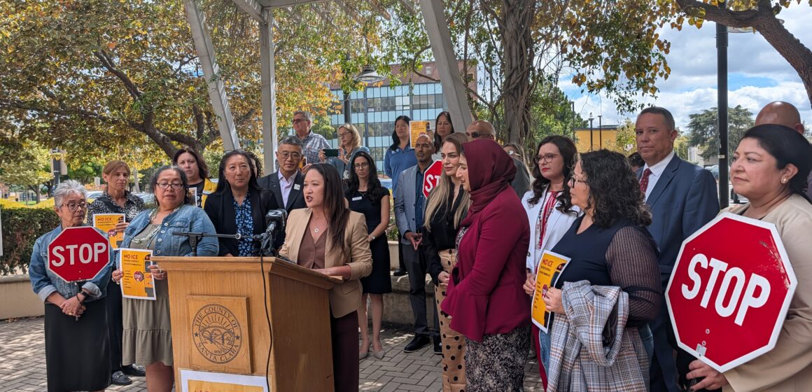 A woman speaking at a podium outside with a crowd of people behind her