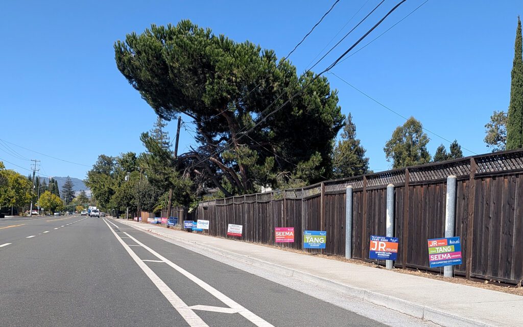 Campaign signs for a local election line a fence along a road in Cupertino, California