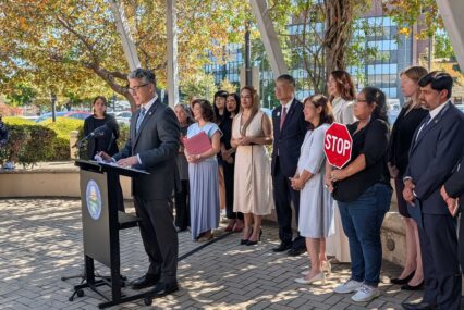 A man speaks at a podium outside with a row of people standing behind him in San Jose, California