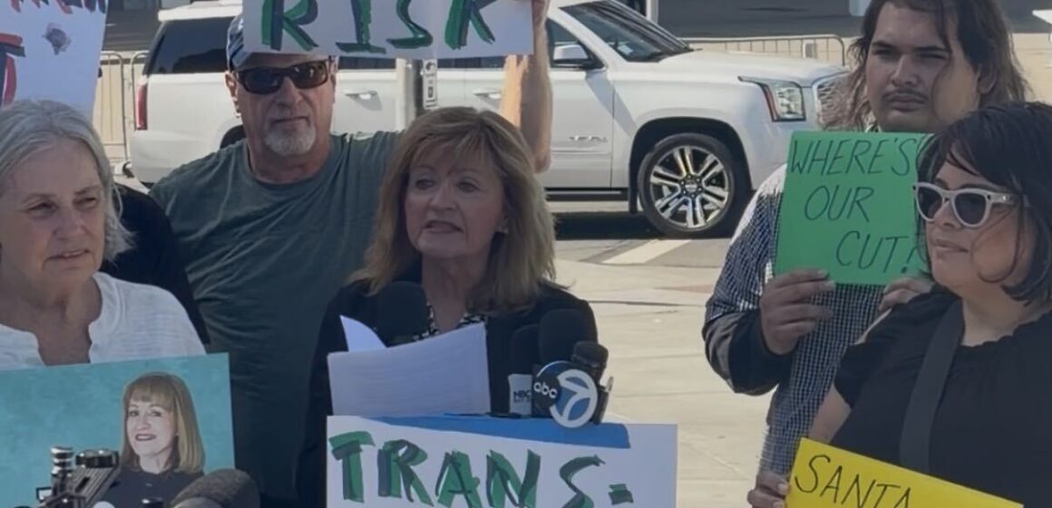 A woman speaking at a podium with people standing behind her holding signs in Santa Clara, California