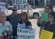 A woman speaking at a podium with people standing behind her holding signs in Santa Clara, California