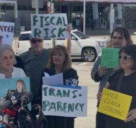 A woman speaking at a podium with people standing behind her holding signs in Santa Clara, California