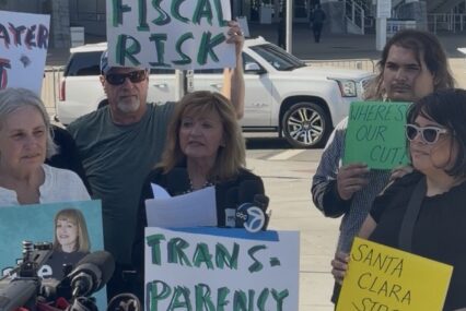 A woman speaking at a podium with people standing behind her holding signs in Santa Clara, California