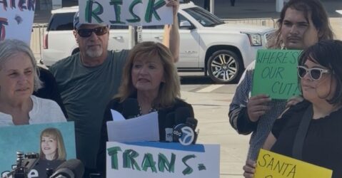 A woman speaking at a podium with people standing behind her holding signs in Santa Clara, California