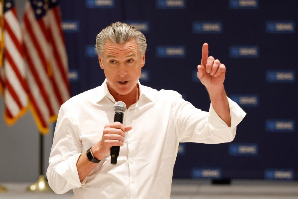 A man in a white button shirt holding a microphone with an American flag in the background